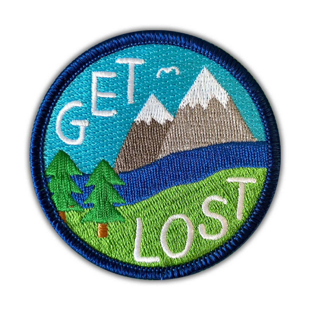 Circular blue and green embroidered patch with snow-capped mountains, pine trees, river, and GET LOST text.